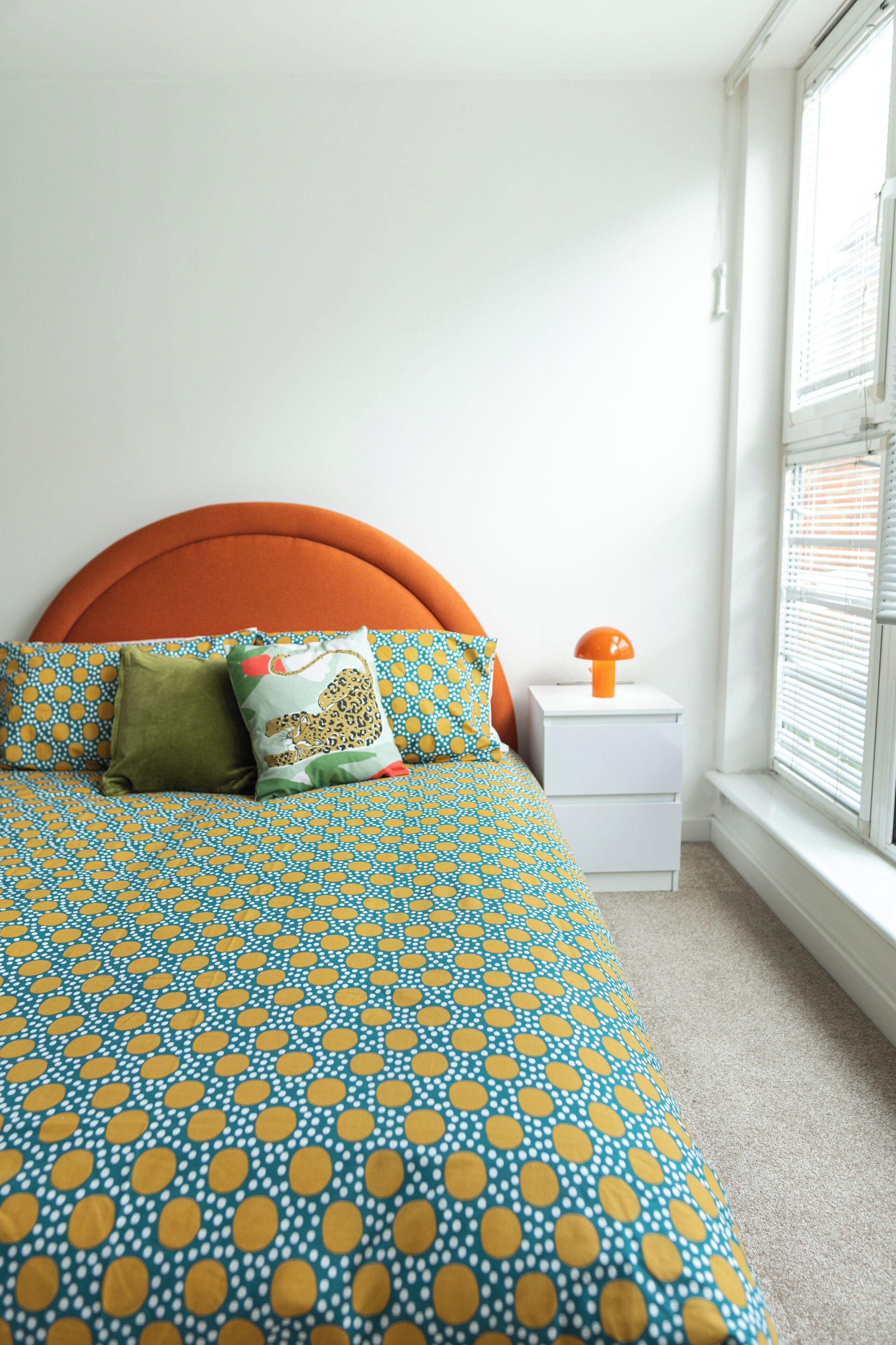 A bed with a patterned bedspread, a nightstand with a lamp, and a window with blinds.