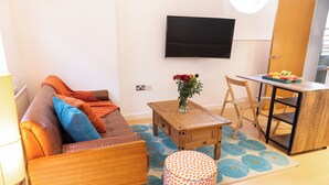 Een moderne woonkamer met een bank, salontafel en poef. Een flatscreen-tv is aan de muur bevestigd, en er staat een eettafel met stoelen.