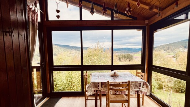 A wooden interior with a table and chairs, large windows offering a view of a forested landscape. - Chalet le Petit Coeur (Xonrupt-Longemer)