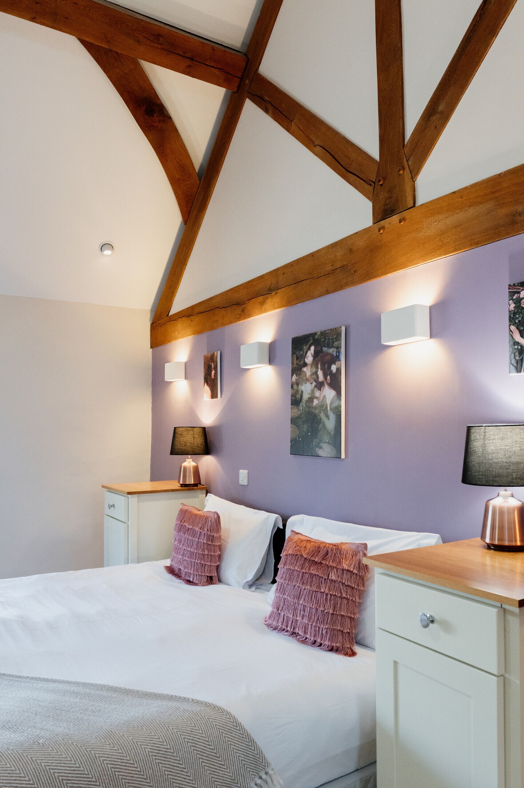 A bedroom with a wooden ceiling, a bed with white bedding, two bedside tables with lamps, and wall-mounted light fixtures.