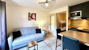 A compact living space with a sofa, a small table, a kitchenette, and a ceiling light fixture. - Le Refuge des Skieurs (Combloux)