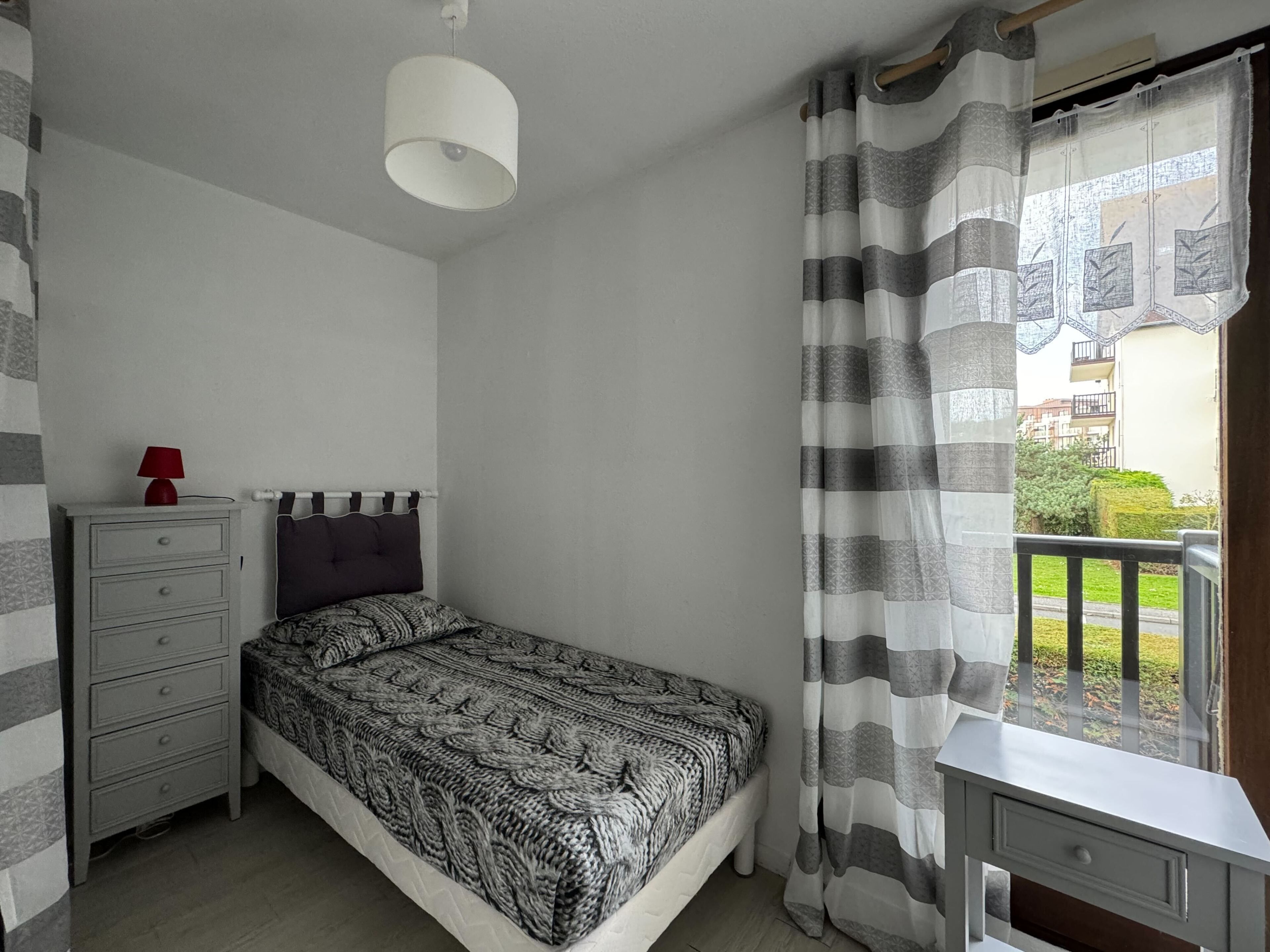 A bedroom with a bed, a nightstand with a lamp, a dresser, and a window with curtains.