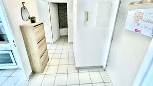 Un pasillo estrecho con suelo de baldosas blancas, un armario de madera, una lámpara colgante, una puerta con un mapa y una planta pequeña sobre el mueble.