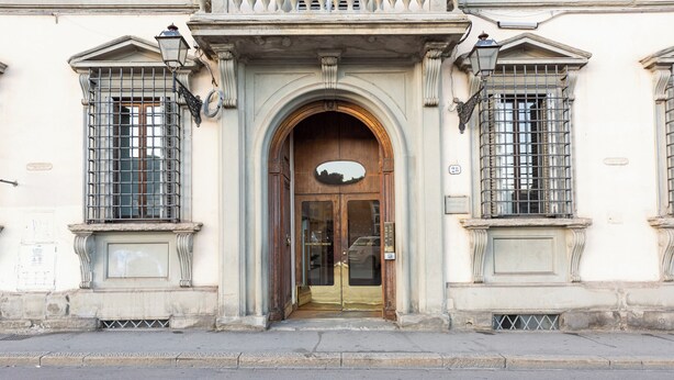 A building facade with a central arched entrance, flanked by two windows with metal bars, and a small balcony above the entrance.