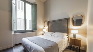 A neatly made bed in a room with a window, a bedside table with a lamp, and a mirror on the wall.
