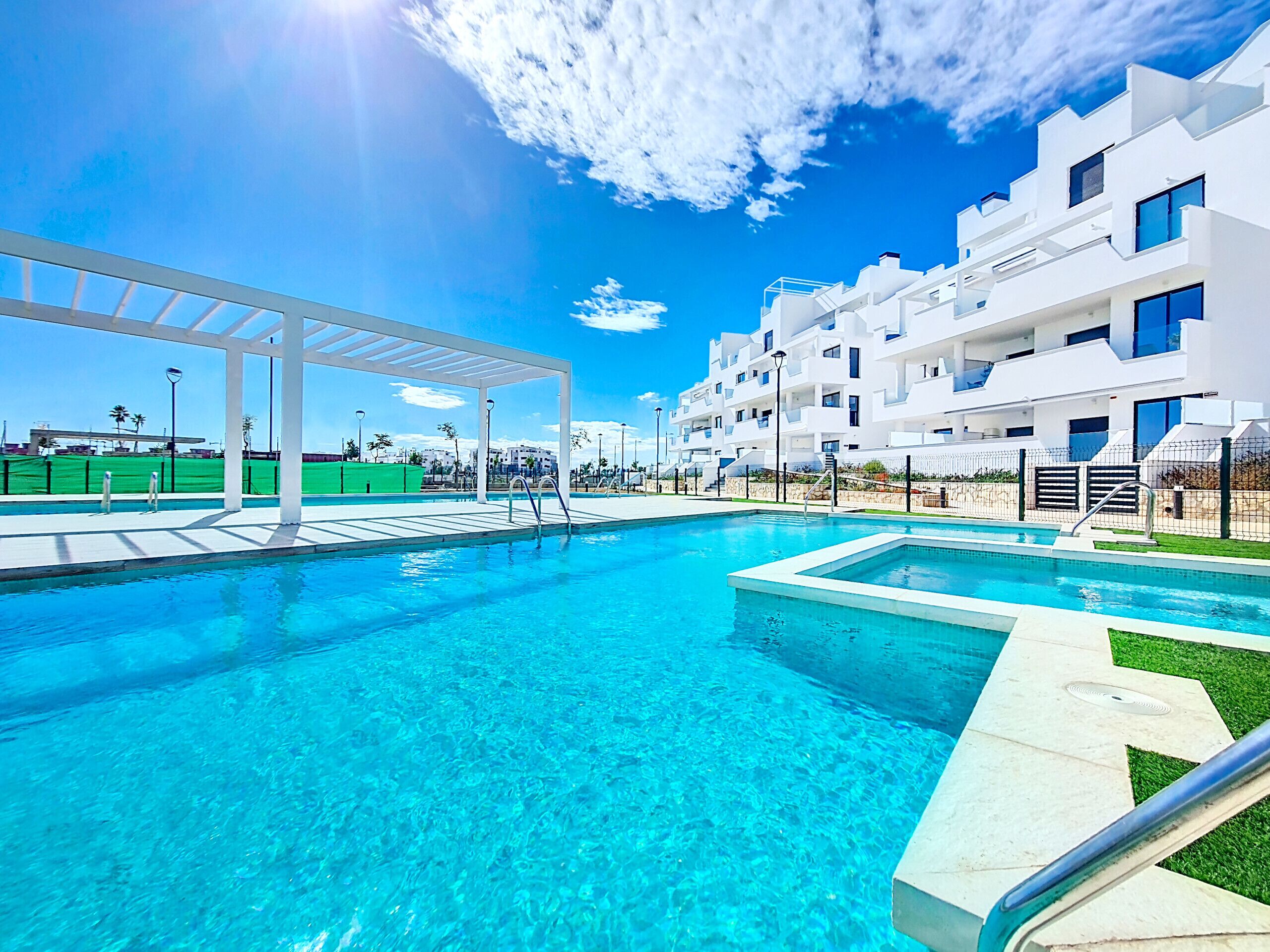 A clear blue pool with a white pergola and modern white buildings in the background.