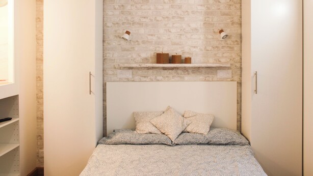 Une chambre avec un mur décoré d’un motif en briques, une étagère avec des bougies et un lit recouvert d’une literie à motifs.