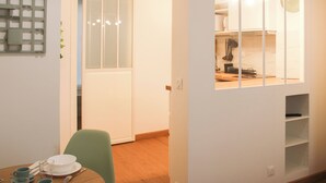 Un coin repas avec une table dressée pour deux personnes, une chaise verte et une cuisine visible à travers une cloison en verre.