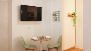 Un petit coin repas avec une table ronde, deux chaises, un téléviseur fixé au mur, un luminaire et une petite plante.