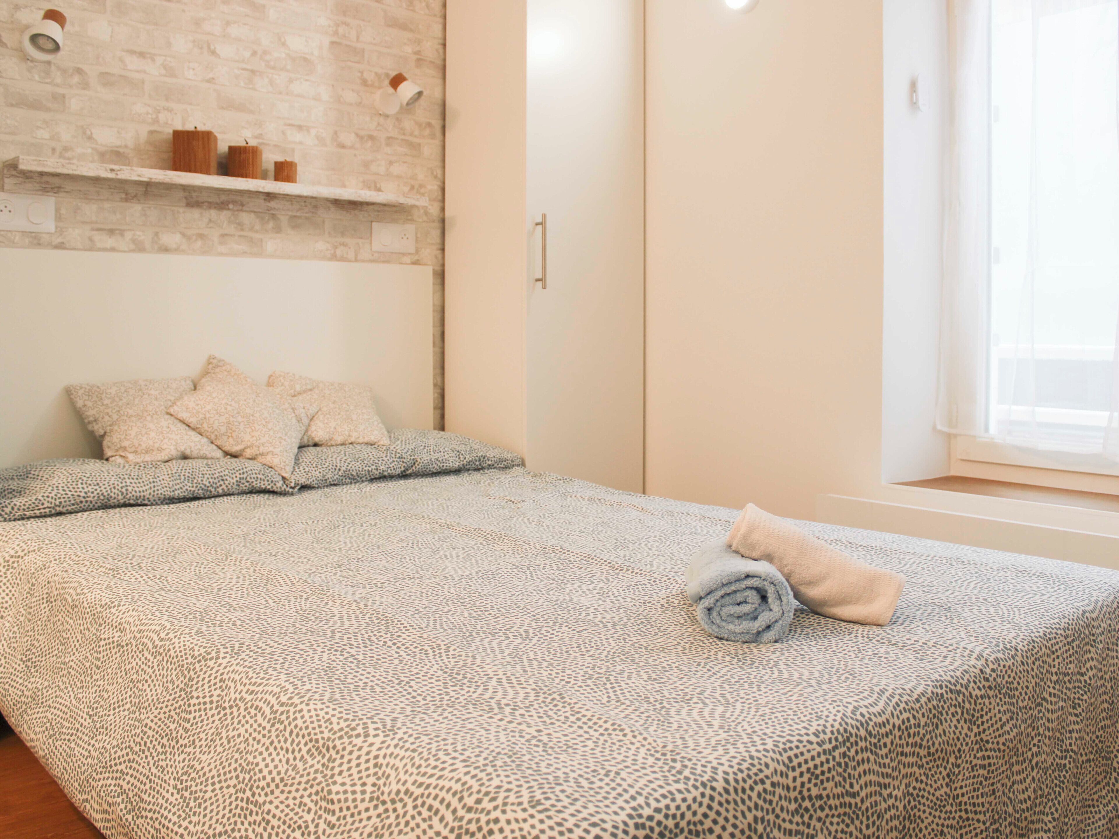 A bedroom with a patterned bedspread, pillows, a rolled towel, a brick wall, a shelf with candles, and a window with curtains.