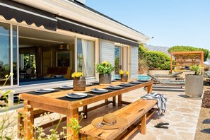 Un área de comedor al aire libre con una mesa de madera dispuesta para cuatro personas, rodeada de plantas en macetas y una zona de descanso junto a la piscina.