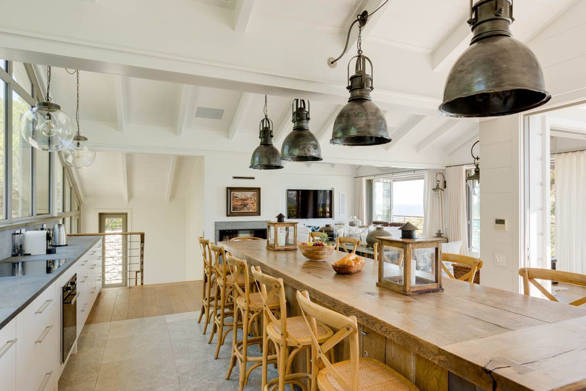 A spacious kitchen with a long wooden bar, hanging industrial-style pendant lights, and a view of the sea through large windows.