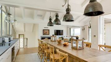 A spacious kitchen with a long wooden bar, hanging industrial-style pendant lights, and a view of the sea through large windows.