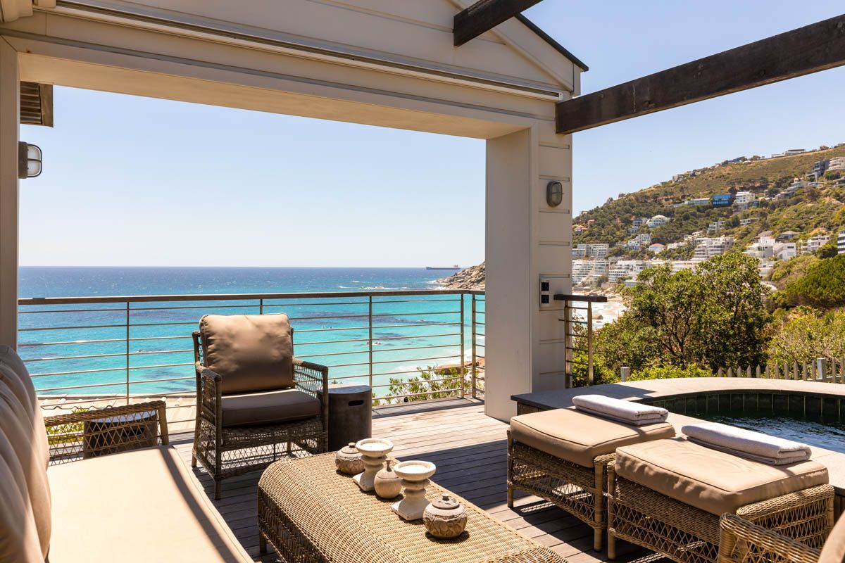 A balcony with wicker furniture, a small pool, and a view of the sea and hillside buildings.