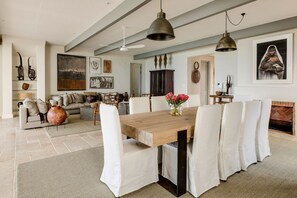 Un vaste espace dédié à la salle à manger, avec une table en bois et des chaises blanches, une cheminée et diverses œuvres d’art accrochées aux murs.
