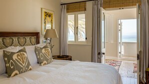 A hotel room with a large bed, a bedside lamp, a painting on the wall, and a view of the ocean through an open door.