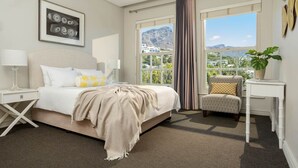Ein Hotelzimmer mit Bett, Nachttischen, einem Sessel, einer Zimmerpflanze und einem Fenster mit Blick auf Berge und Gebäude.