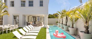 Ein Pool mit Liegestühlen und einem Flamingo-Schwimmding, umgeben von Topfpflanzen und einem weißen Gebäude.