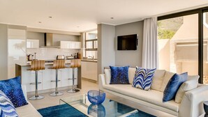 Una sala de estar moderna con un sofá, una mesa de centro de vidrio y una zona de cocina con taburetes de bar.