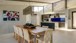 Una cocina moderna con una isla grande, un comedor con sillas de mimbre y vistas a montañas a través de amplios ventanales.