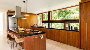 Eine moderne Küche mit Holzschränken, einer zentralen Kochinsel mit Barhocker und großen Fenstern mit Blick ins Grüne.