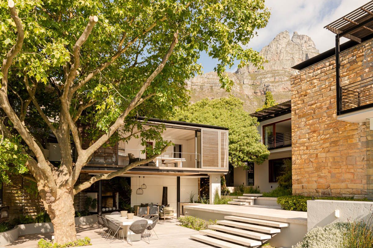 A modern two-story house with a patio area, outdoor furniture, and a large tree.