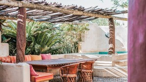 Un patio al aire libre con una mesa y sillas de madera, rodeado de abundante vegetación y un muro de piedra.