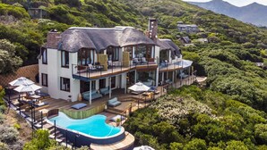 Une grande maison à plusieurs étages, dotée d’une piscine, entourée d’une végétation luxuriante et de collines.