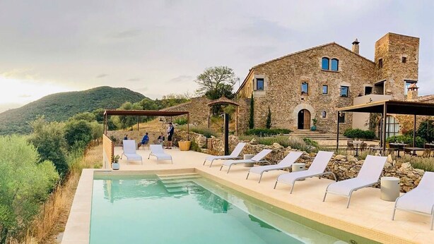 Zona de piscina con sillones de descanso y un edificio de piedra al fondo.