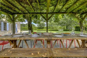 An outdoor dining area with a wooden table set for a meal, surrounded by trees and a stone building.