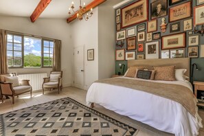 A bedroom with a large bed, a patterned rug, a wooden chair, a window with curtains, and a wall adorned with framed pictures.