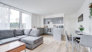 A modern living space with a grey sectional sofa, a dining area with a table and chairs, a kitchen with grey cabinets and appliances, and a large window providing natural light. - Cozy London Apartment With 2 Bedrooms (London)