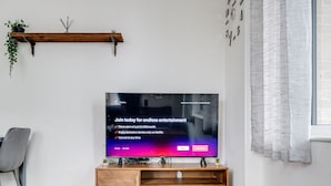 A modern living room with a flat-screen TV on a wooden stand, a grey chair, and a plant on a shelf. - Cozy London Apartment With 2 Bedrooms (London)