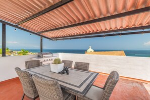 A covered outdoor dining area with a view of the ocean, a grill, wicker chairs, and a potted plant.