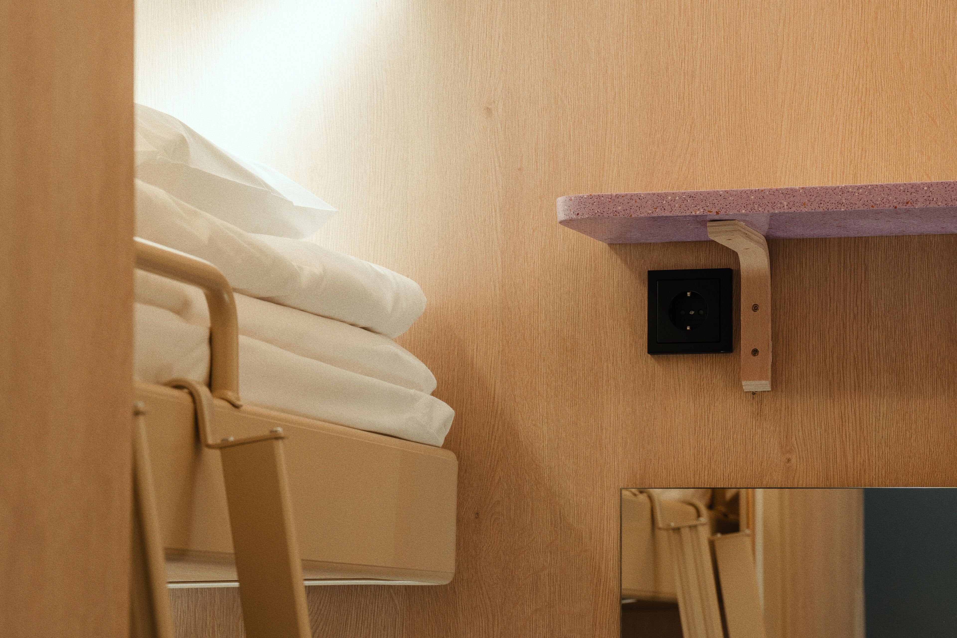 A wooden wall with a shelf, a bed with white linens, and a small black device on the wall.