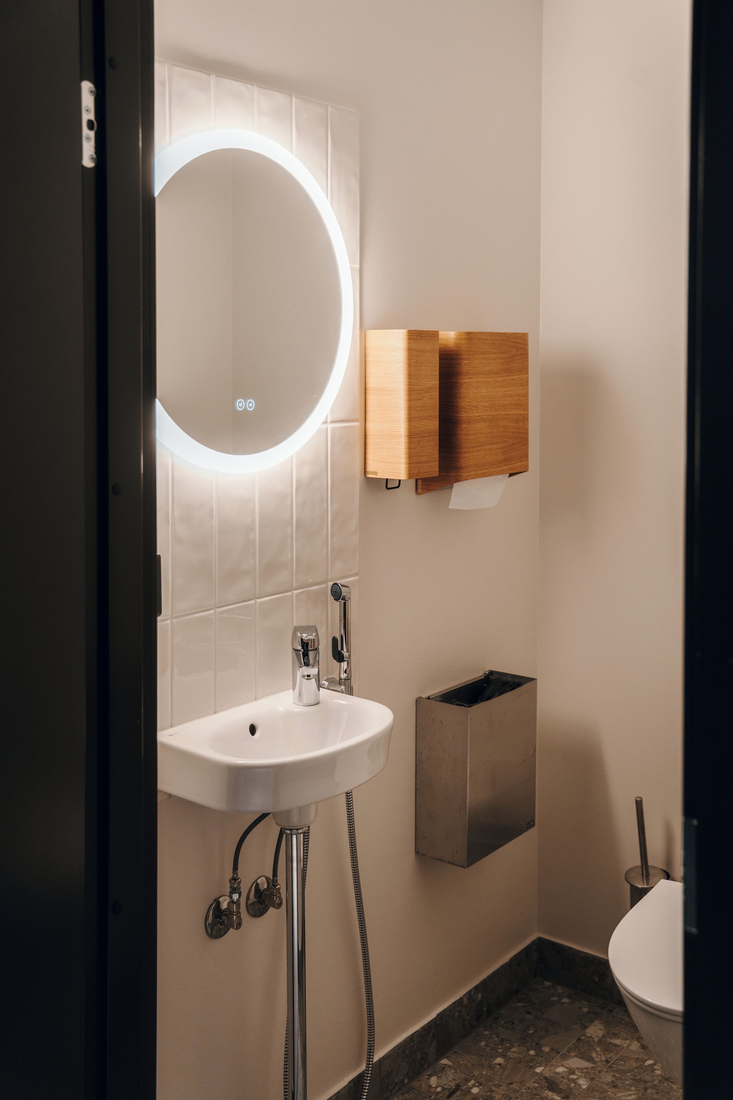 A modern bathroom with a white sink, a round mirror with a light, a wooden cabinet, and a toilet.