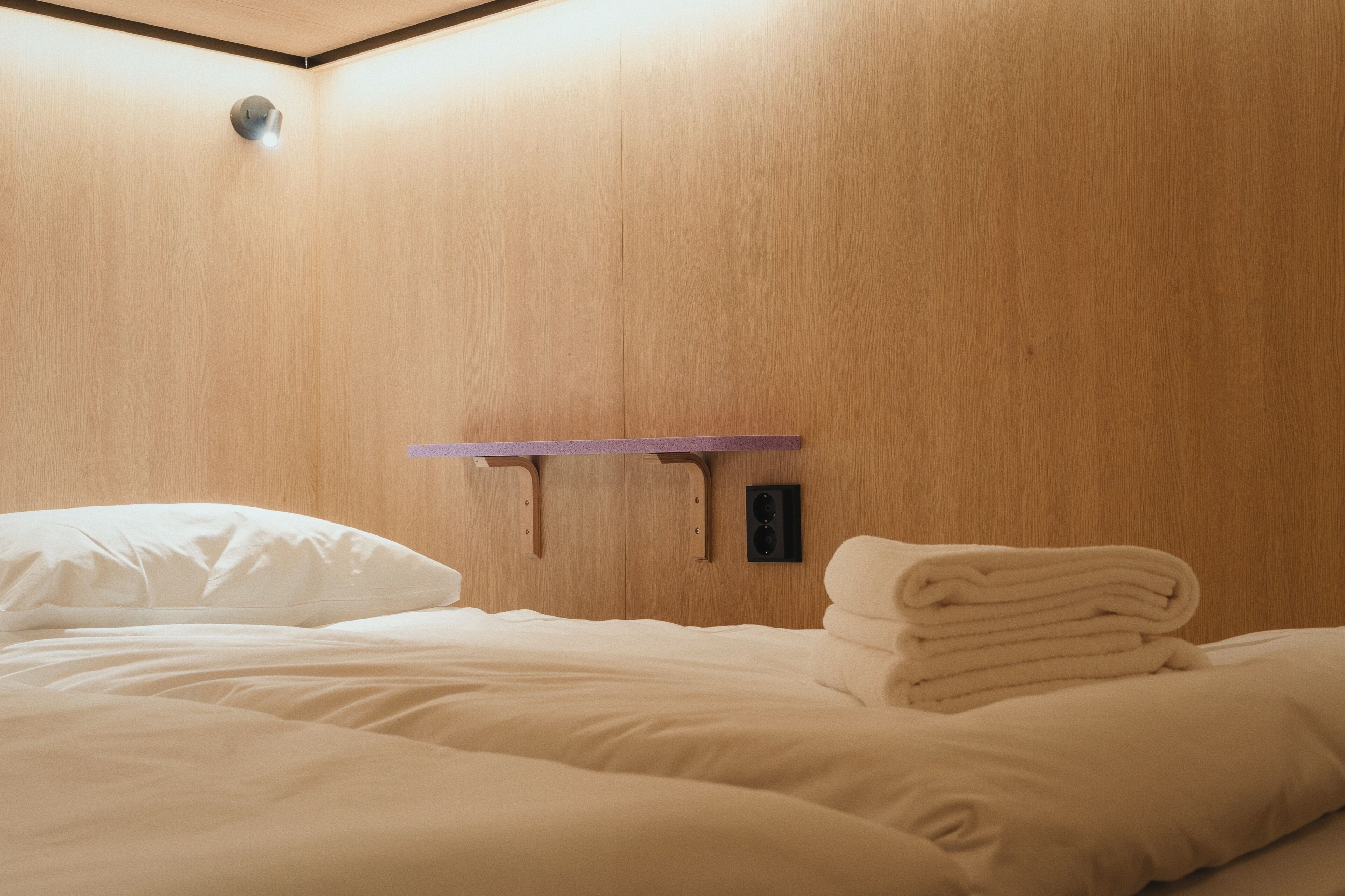 A neatly made bed with white linens and a stack of folded towels on a wooden wall.