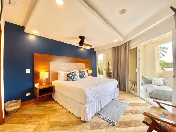 Ein Schlafzimmer mit einem großen Bett, einem Deckenventilator, einem Nachttisch und Blick auf den Balkon.