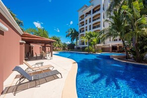 Ein Poolbereich eines Resorts mit Liegestühlen und einer überdachten Sitzgelegenheit, umgeben von Palmen und einem mehrstöckigen Gebäude.