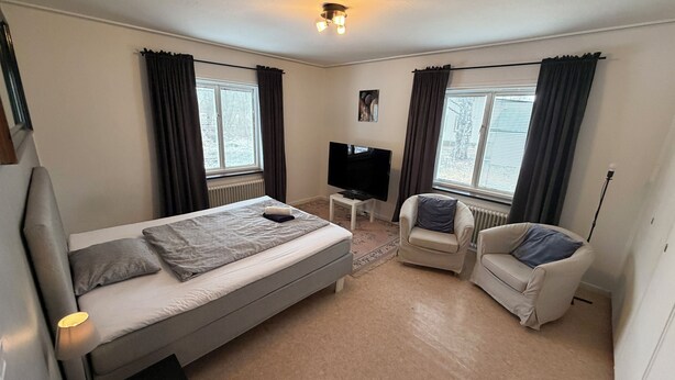 A bedroom with a bed, a TV, a chair, and two windows with curtains.