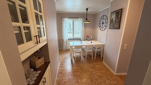 A narrow hallway with a dining area featuring a table and chairs, a clock on the wall, and a window with blinds.