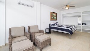 Une chambre à coucher comprenant un lit, deux fauteuils en osier, un ventilateur de plafond, un tableau et des étagères intégrées.