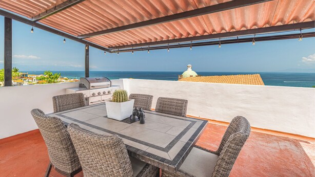 Un espace extérieur abrité, aménagé pour les repas, avec vue sur l’océan, un barbecue, des chaises en osier et une plante en pot.