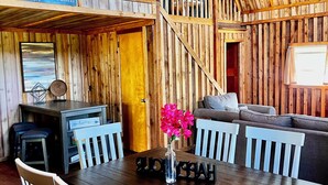 Interior de una cabaña con una mesa de comedor de madera, sillas, un sofá y una escalera que sube a un nivel superior.