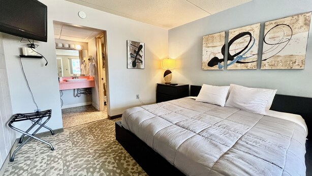 A hotel room with a large bed, a bedside table with a lamp, a TV mounted on the wall, and a bathroom visible through an open door. - Downtown CF Studio - Free Gated Parking! 516 (Cedar Falls)