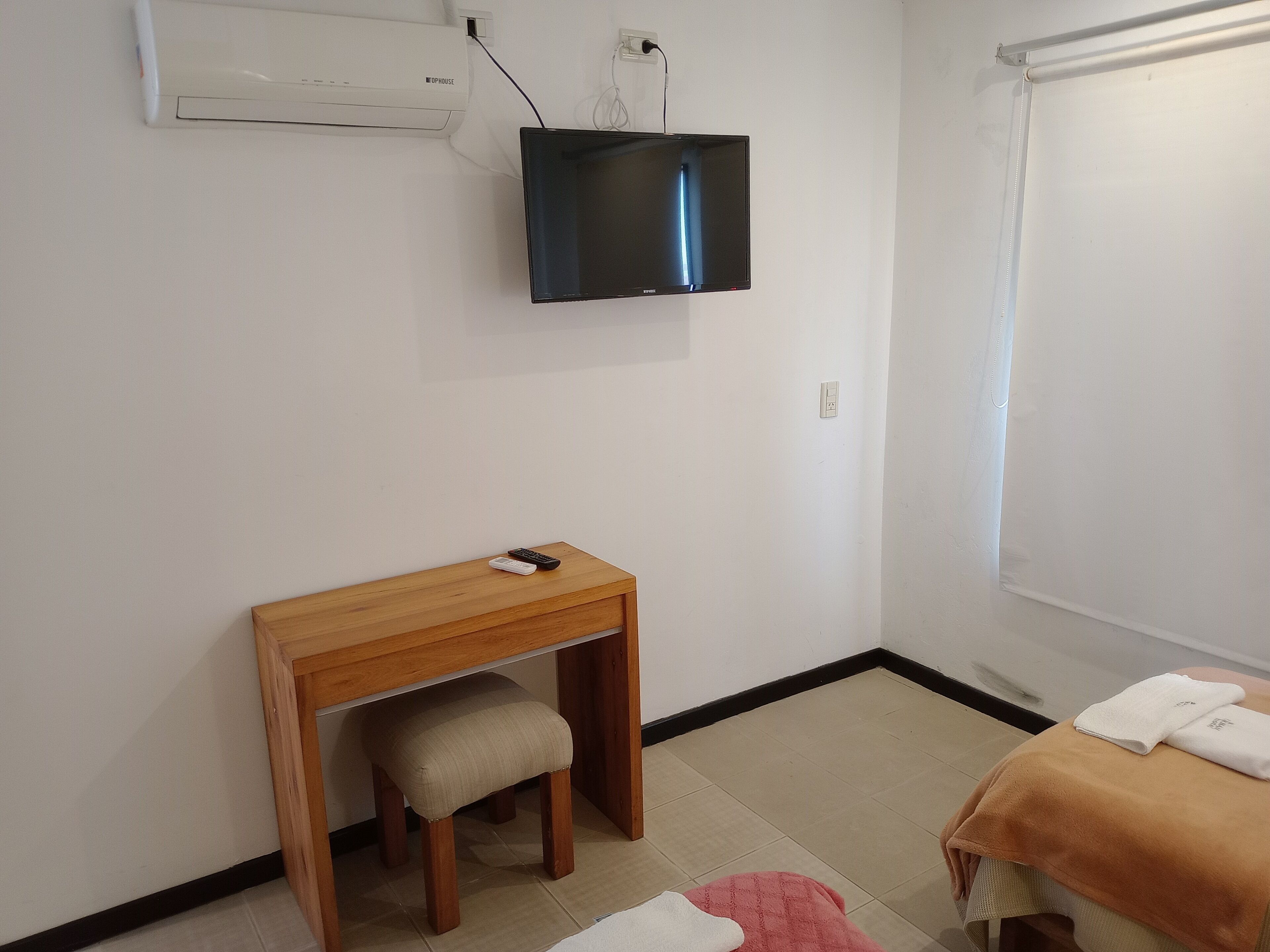 Una habitación pequeña con un escritorio de madera, un televisor montado en la pared, un taburete y una cama con una toalla.