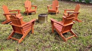 Een overdekt terras met vier houten Adirondack-stoelen en een vuurkuil in het midden, omgeven door een gazon en bomen.