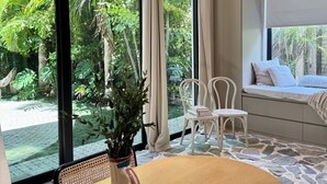 A room with a large window, a wooden table, a white chair, and a sofa with a view of greenery outside. - Atelier Lumi - @_lumicollection (Miami)