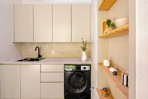 A compact kitchen with a washing machine, a sink, and a small shelf with a vase and a mug. - Atelier Lumi @_lumicollection (Miami)