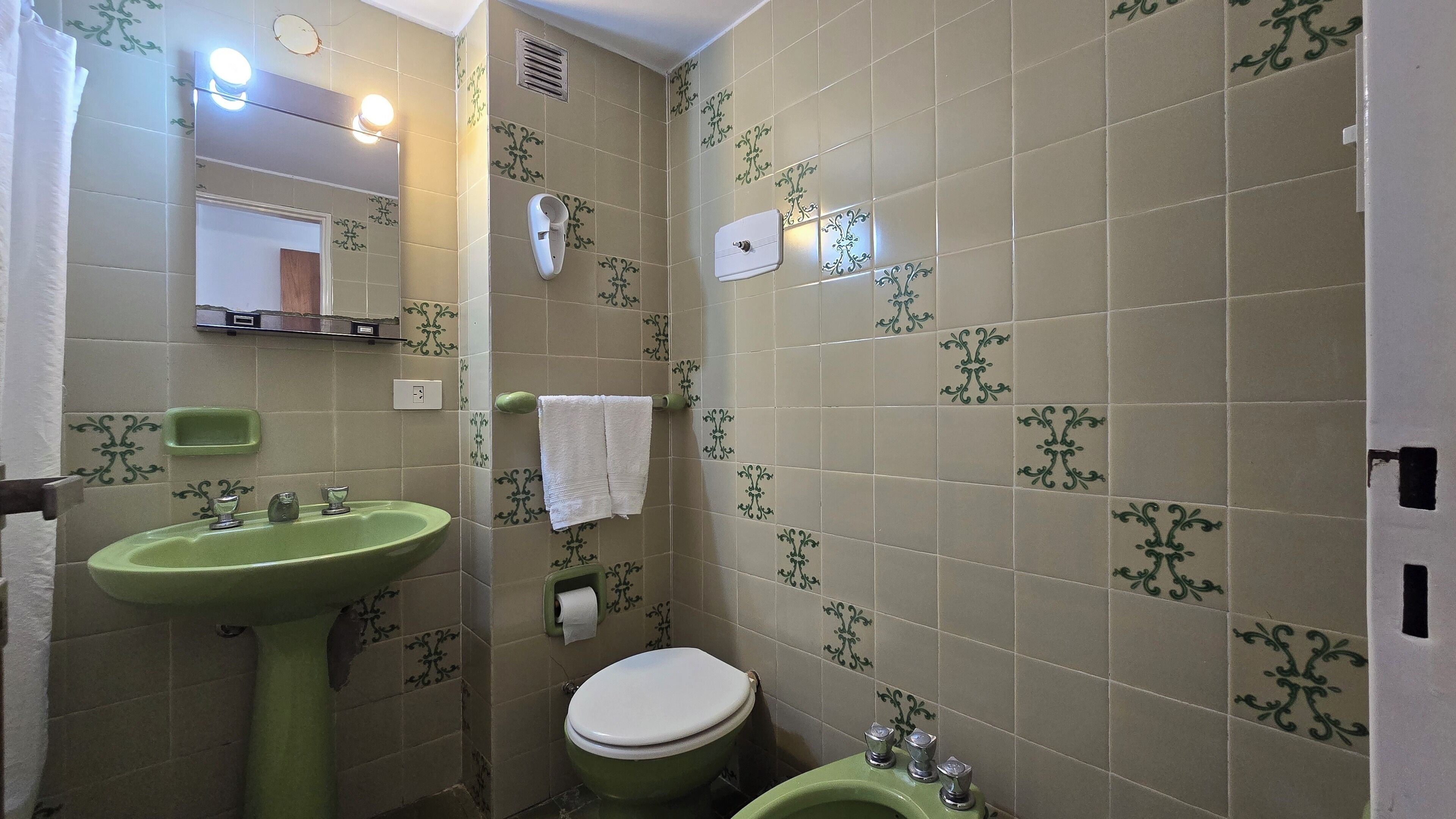 A bathroom with a green sink, a toilet, and a mirror. There is a towel hanging on the wall and a soap dispenser.